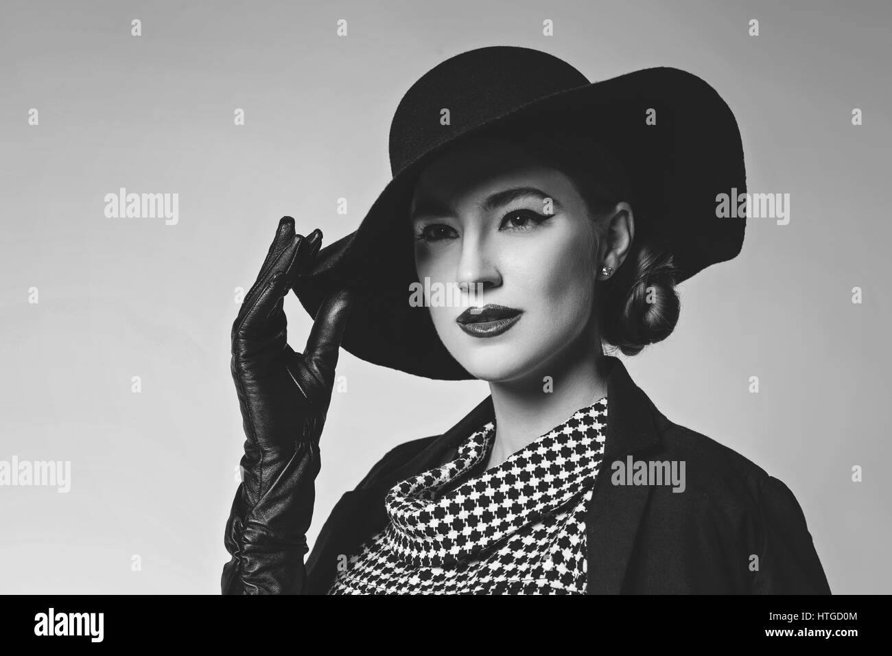 Bella giovane donna con labbra rosse per il make-up retrò in bianco e nero vestito, cappello, giacca e guanti in pelle in piedi su sfondo grigio. Monocromatico. Foto Stock