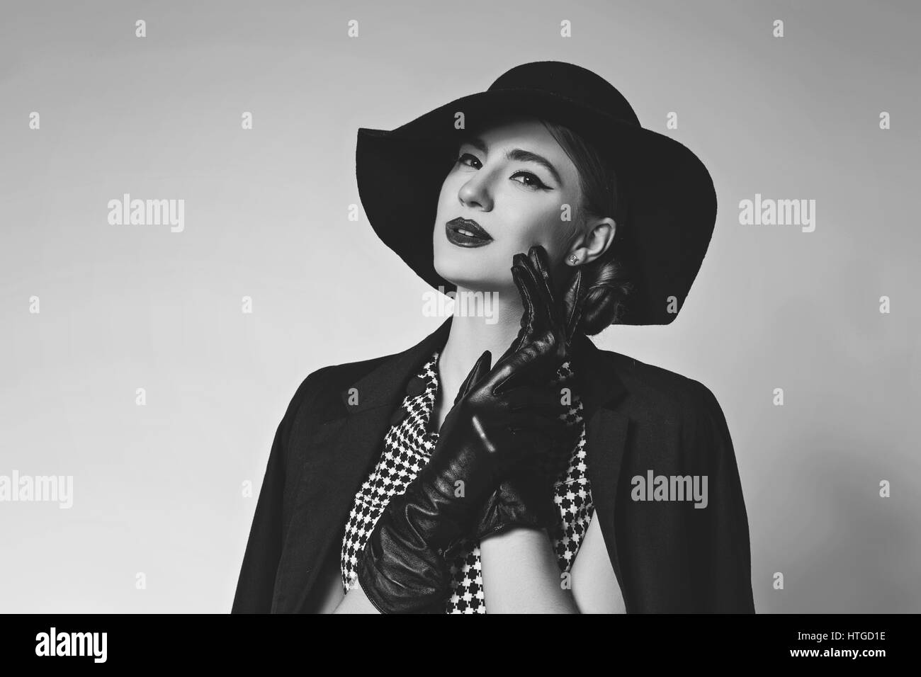 Bella giovane donna con labbra rosse per il make-up retrò in bianco e nero vestito, cappello, giacca e guanti in pelle in piedi su sfondo grigio. Monocromatico. Co Foto Stock