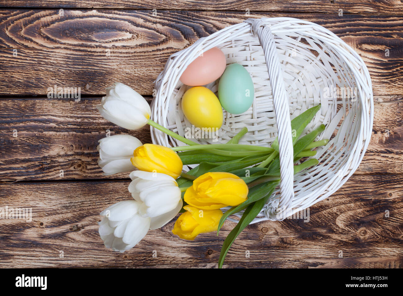 Uova di Pasqua in un cesto bianco con giallo e tulipani bianco su sfondo di legno Foto Stock