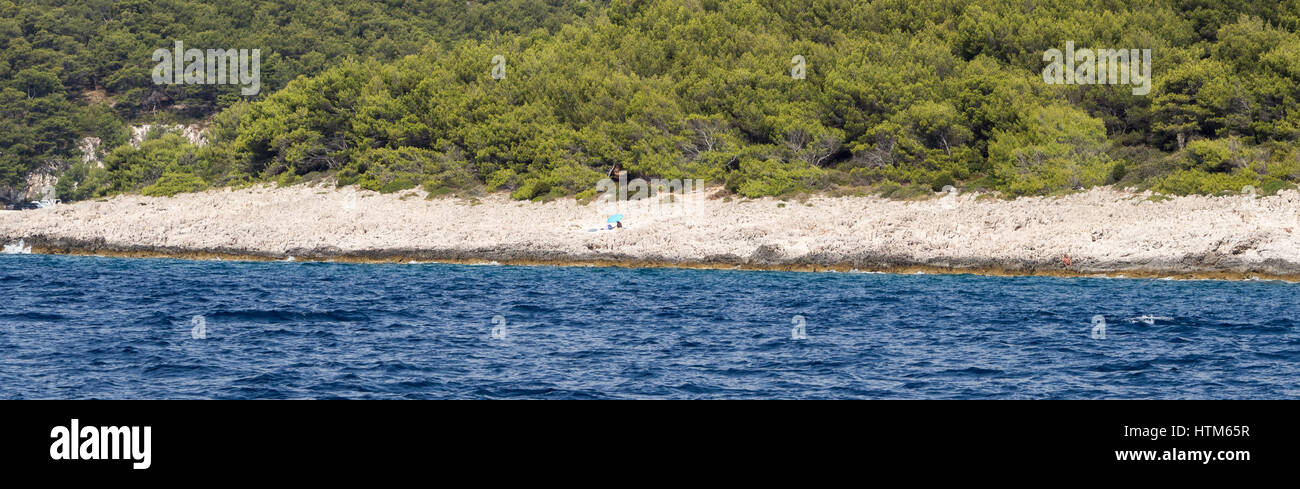 Un lone unico sunbather da soli su un isolato sassoso shingle spiaggia rocciosa sotto l'ombra di un ombrellone blu ombrello in Croazia del Sud Foto Stock