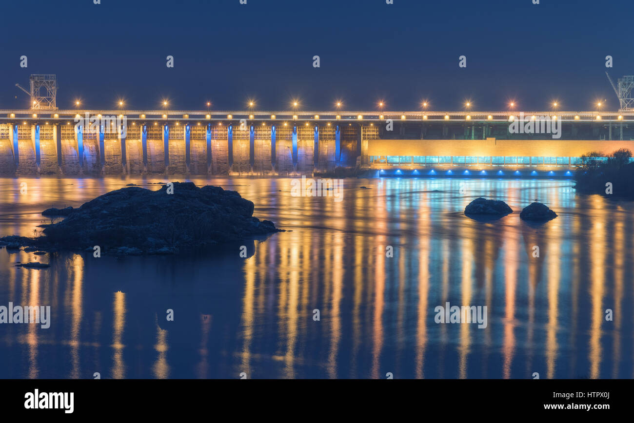 Diga di notte. Bellissimo paesaggio industriale con la diga idroelettrica, il ponte sul fiume, illuminazione della città riflessa nell'acqua, rocce e cielo. Foto Stock