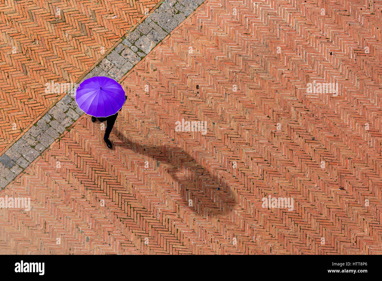 Anonimo man in black suit è a piedi in rosso e marrone quadrato di piastrelle in Italia, nascondendo dal sole con un ombrello viola. La sua ombra potrebbe essere visto. Foto Stock