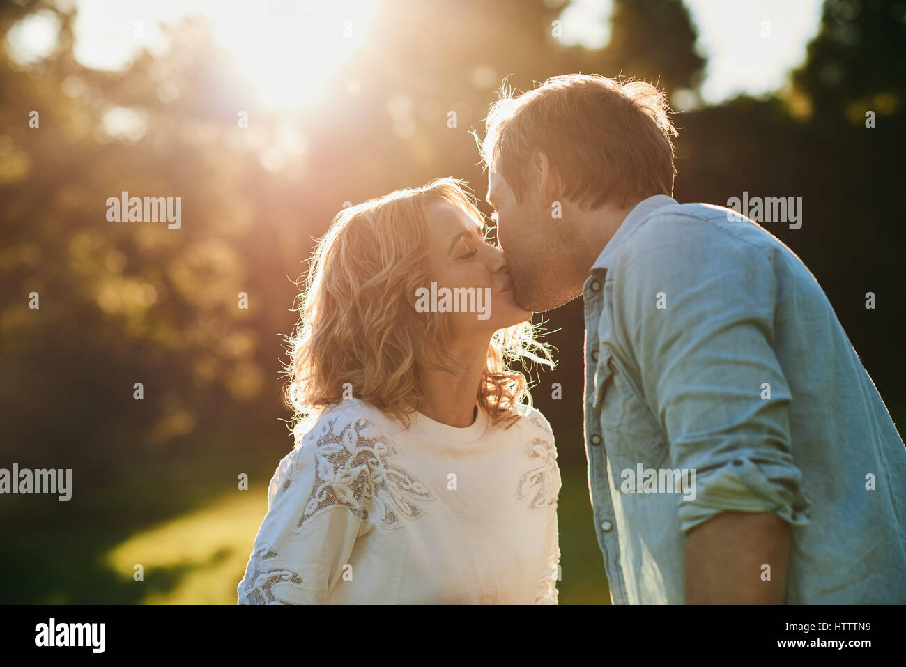 Affettuosa coppia giovane kissing ogni altro con gli occhi chiusi mentre godendo un romantico estivo soleggiato pomeriggio insieme in un parco Foto Stock