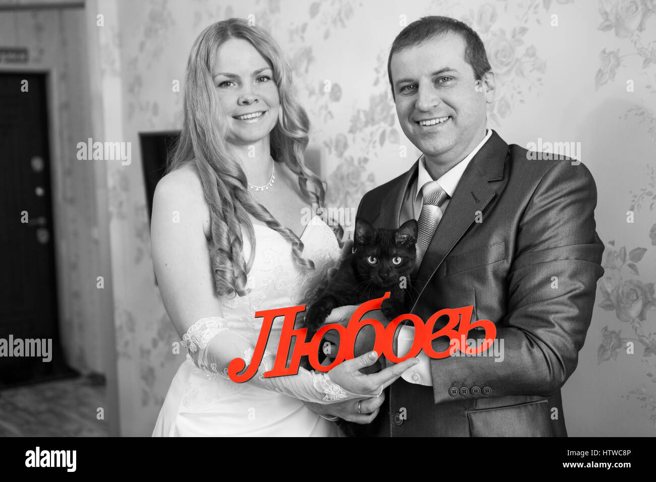 Caucasian sposa e lo sposo con gatto nero e la parola amore in russo in mani. Stanza vuota, inaugurazione della casa nuova. Immagine in bianco e nero Foto Stock