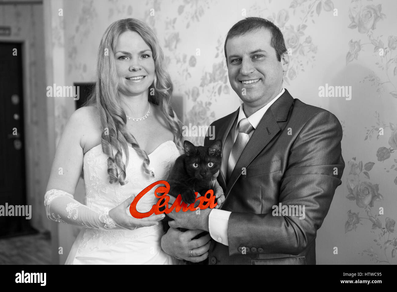 Felice e sorridente Caucasian giovane azienda red parola famiglia in russo in mani e gatto nero, immagine in scala di grigi Foto Stock