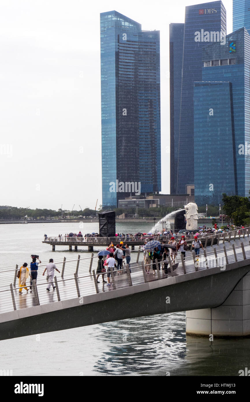La gente camminare attraverso il Ponte del Giubileo, Merlion sgorga acqua in Marina Bay e la baia di Marina Centro finanziario in background. Foto Stock