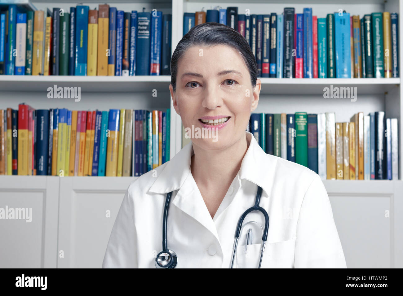 Gentile Dottore con uno stetoscopio davanti a un sacco di libri nel suo ufficio di chirurgia parlando a un paziente, vista frontale, consultazione medica nozione Foto Stock