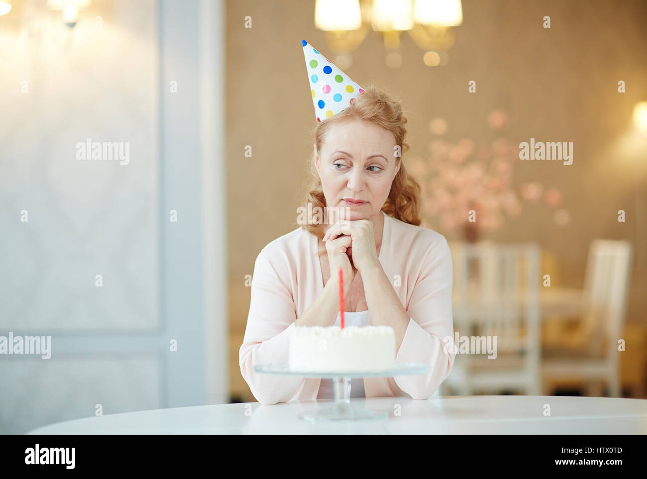 Ritratto di malinconico donna matura seduto da solo al tavolo di compleanno con torta , indossando cappellino e guardando triste Foto Stock