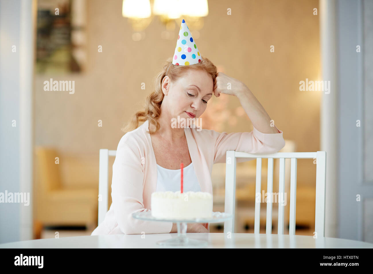 Ritratto di triste lonely donna matura seduto da solo al tavolo di compleanno con torta, indossando il cappellino e attesa per gli ospiti Foto Stock