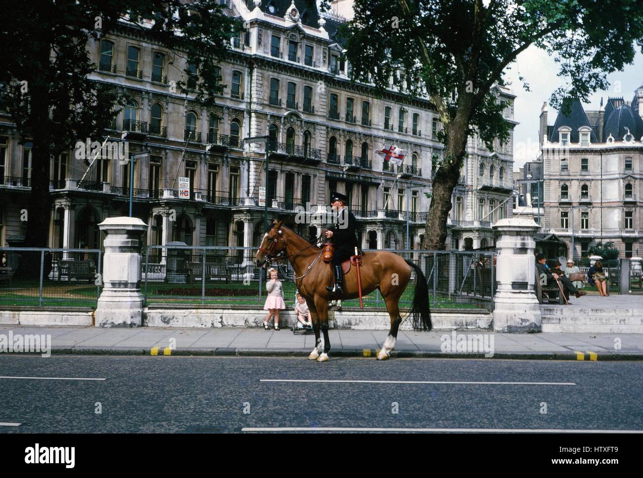 Un poliziotto montato in un nero uniforme e tappo, con una grande billy club, si siede su un cavallo su una strada con un raffinato edificio in background, come due ragazze in abiti rosa a guardare, Regno Unito, 1975. Foto Stock