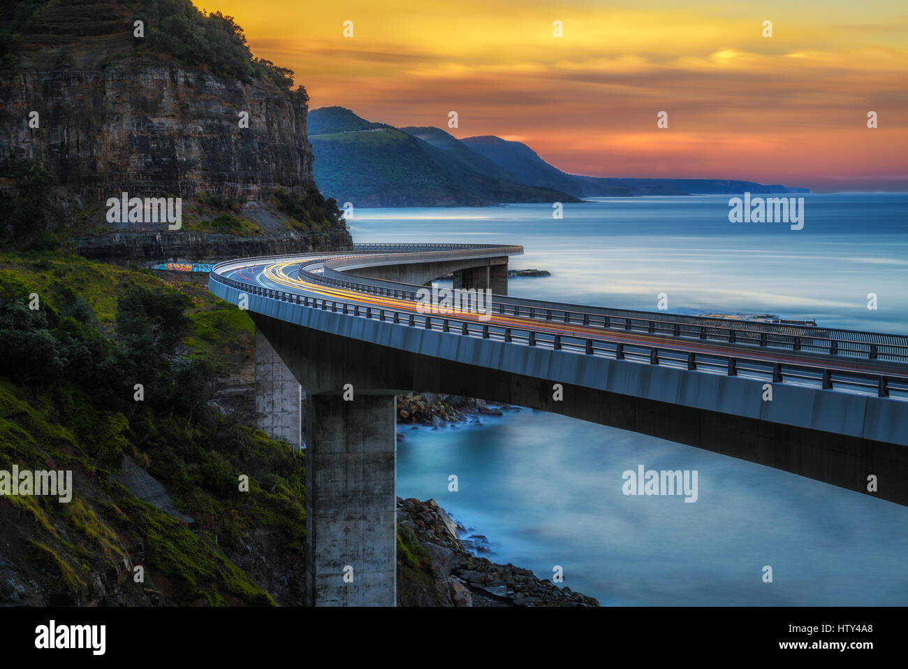 Tramonto sulla scogliera sul mare ponte lungo Australian costa dell'Oceano Pacifico con luci di passaggio di vetture vicino a Sydney, Australia. Lunga esposizione. Foto Stock
