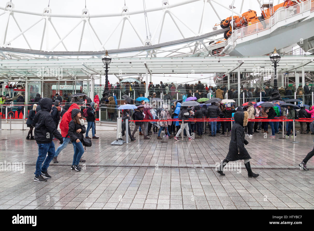 La folla di persone in coda sotto la pioggia battente con ombrelloni in attesa di bordo del London Eye, London, Regno Unito Foto Stock