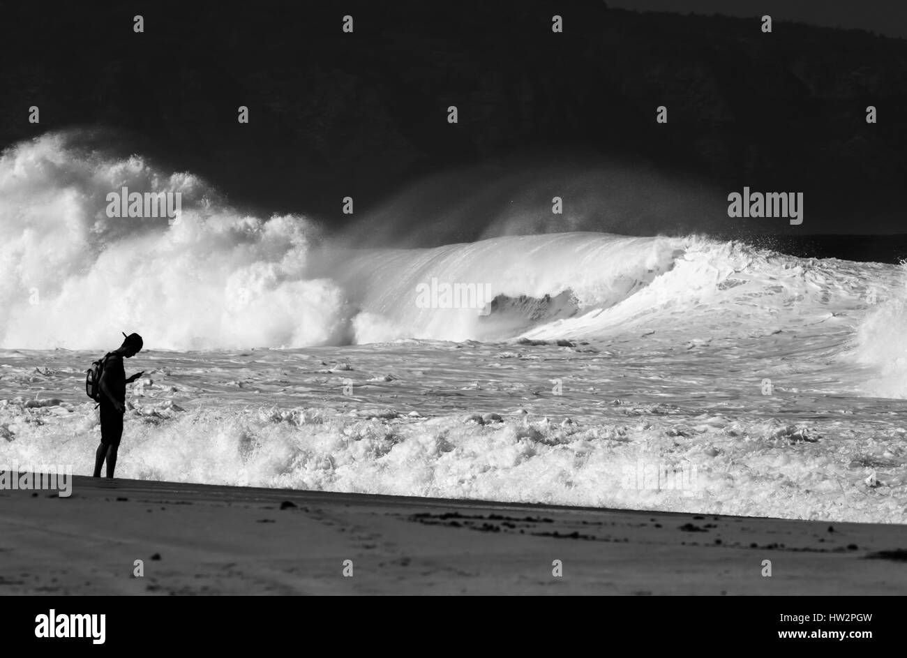 Un giovane uomo controlla il suo telefono con una gigantesca onda di rottura in background, North Shore Oahu Hi Foto Stock