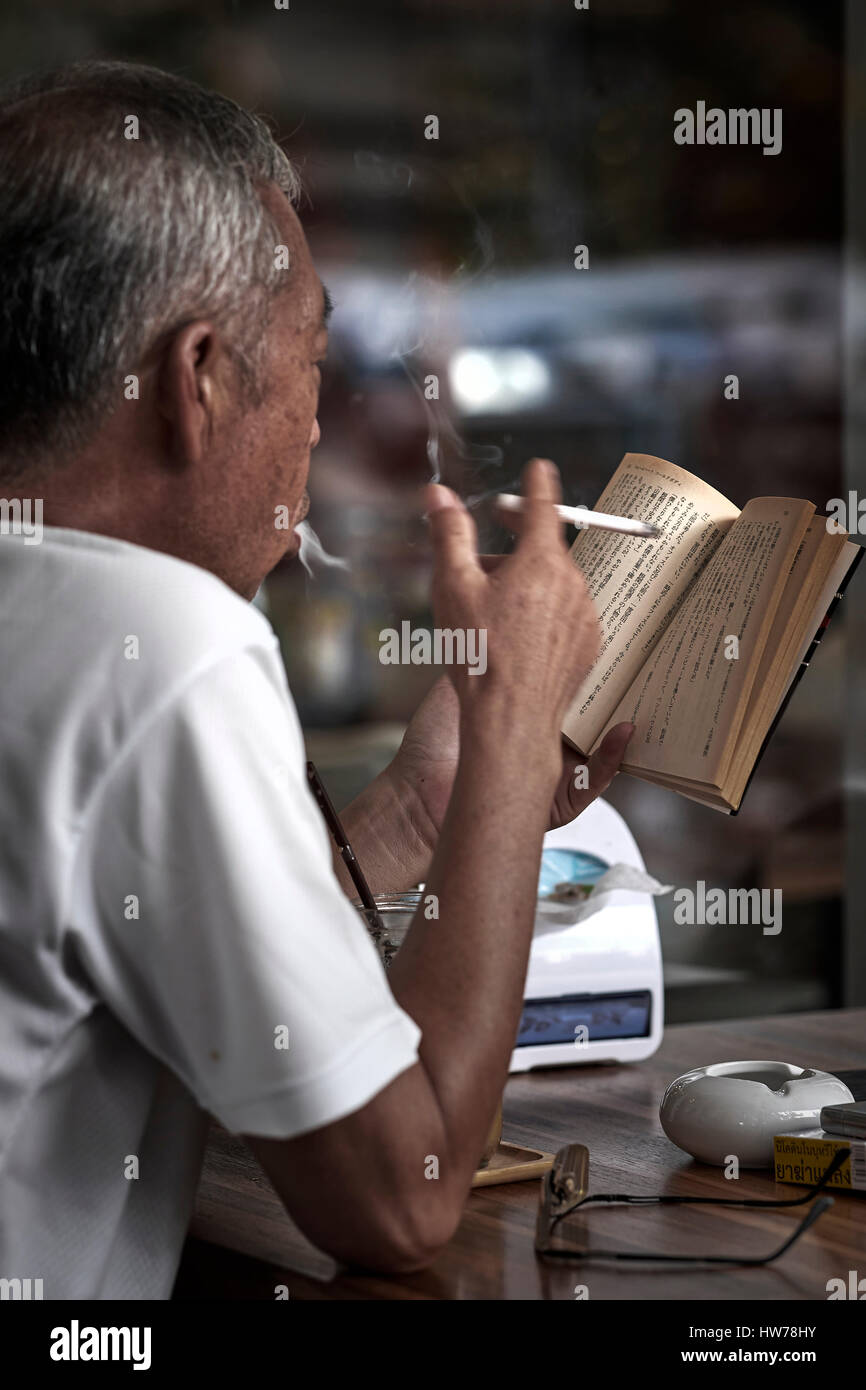 Libro di lettura cinese. Uomo cinese anziano che fuma una sigaretta e legge un libro con iscrizione cinese Foto Stock