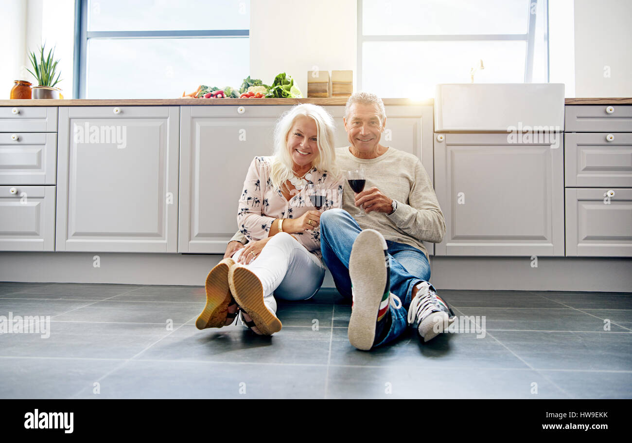 Ridendo Coppia matura azienda vino in cucina mentre si è seduti sul pavimento piastrellato. Copiare lo spazio intorno a loro. Foto Stock