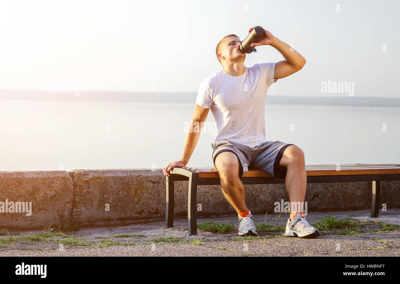 Giovane forte uomo caucasico con cuffie bianche beve un cocktail di proteine da un agitatore dopo aver eseguito all'aria aperta. Il sole luminoso si accende. Foto Stock