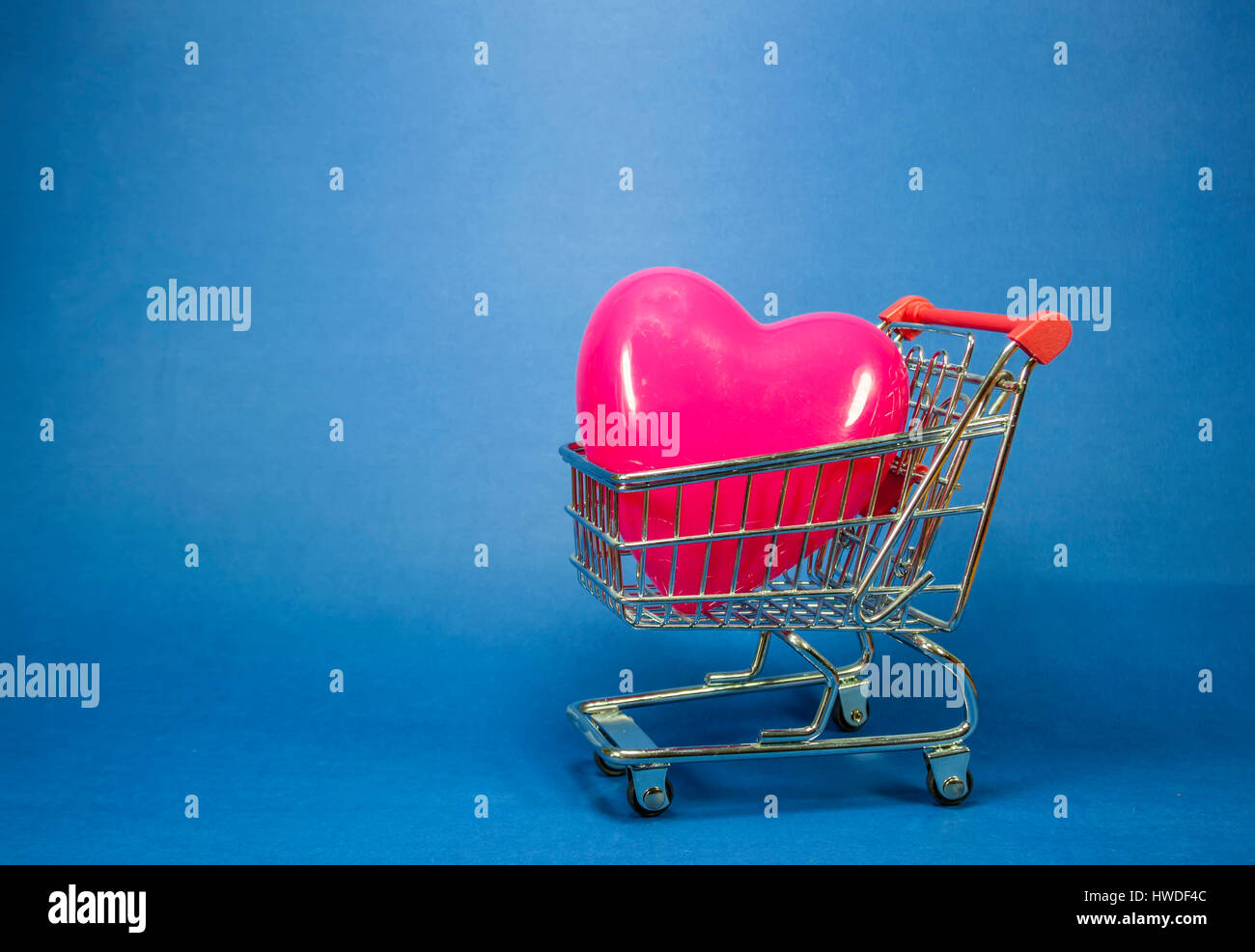 Cuore in plastica rosso di forma all'interno di una miniatura del carrello su sfondo blu, immagine concettuale circa l'amore come una merce nella società consumistica Foto Stock