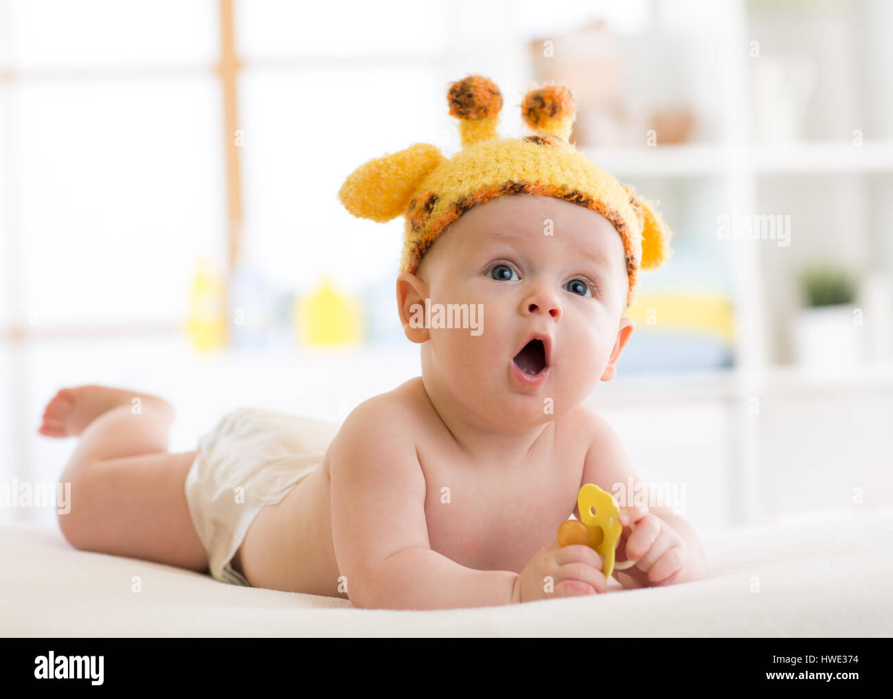 Funny baby boy in giraffa hat sdraiato sul suo ventre in vivaio. Ragazzino che giace sul letto con la bocca aperta e detiene il succhietto in mani Foto Stock