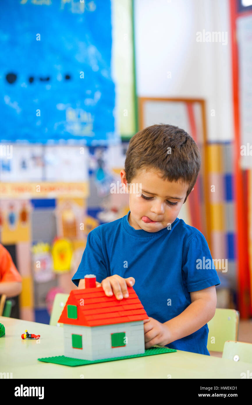 Ragazzo di età prescolare con la lingua di fuori la costruzione di un blocco di giocattolo casa in aula Foto Stock