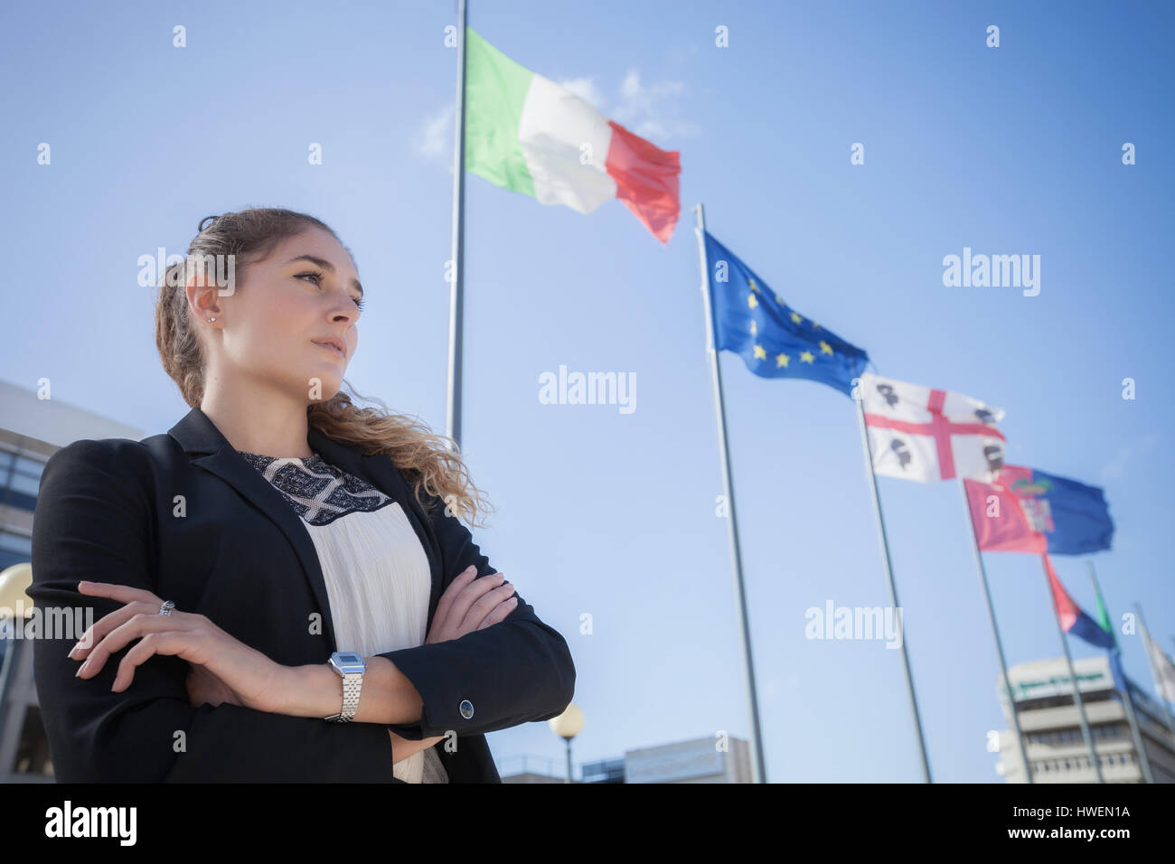 Ritratto di giovane imprenditrice, all'aperto, braccia incrociate, bandiere europee battenti in background Foto Stock