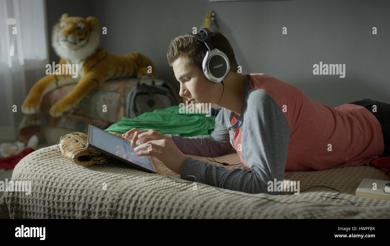 Ragazzo adolescente ascoltando le cuffie e con tavoletta digitale sul letto Foto Stock