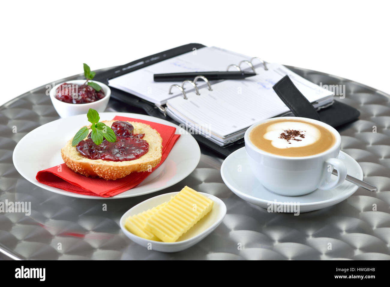 La prima colazione business comprende: un nuovo rotolo di sesamo con marmellata di ciliege e una tazza di cappuccino; un personal organizer in background Foto Stock