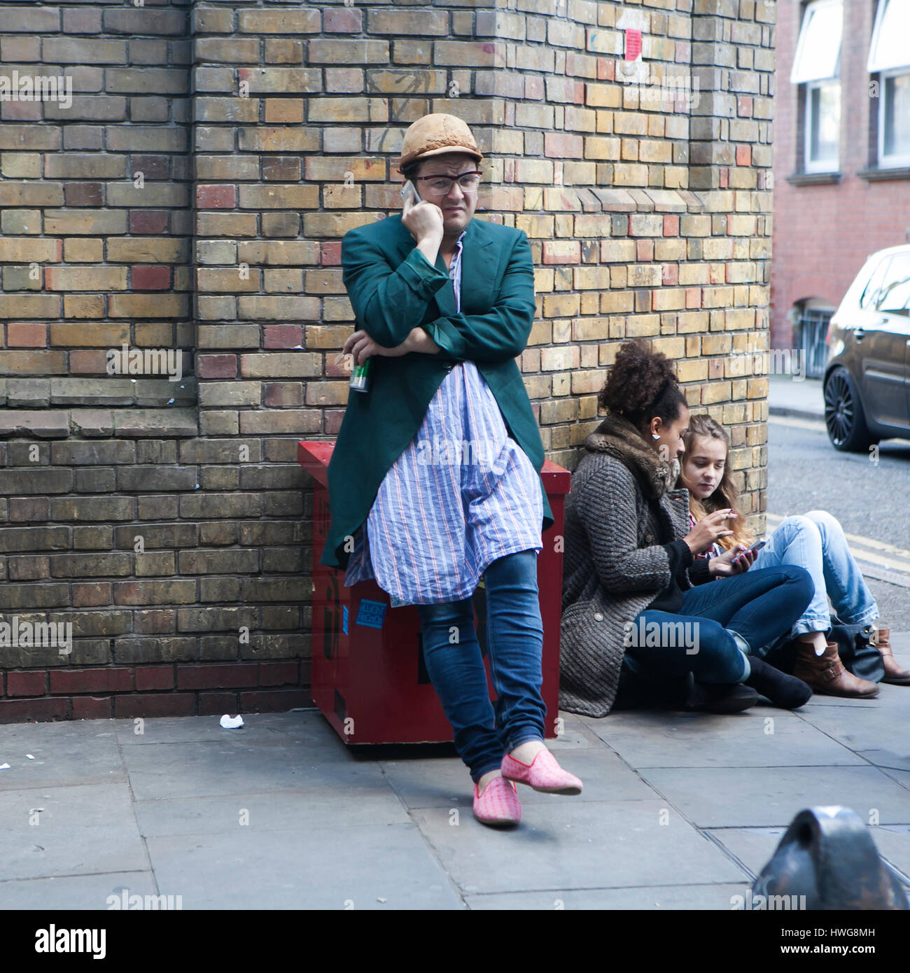 Londra, Inghilterra - Luglio 12, 2016 Un uomo in giacca verde e beige cap sta parlando al telefono, appoggiata contro il muro. Egli si presenta come un tipico offrono Foto Stock