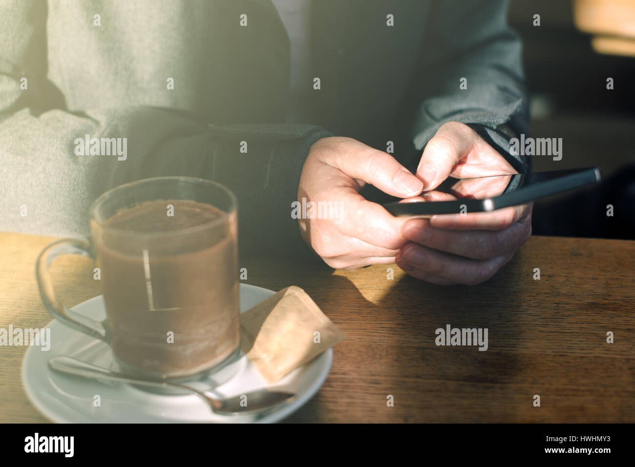 Donna che utilizza il telefono cellulare e di bere cioccolata calda in cafe. Casual giovane adulto femmina caucasica dietro il vetro la digitazione di un messaggio di testo sullo smartphone ap Foto Stock