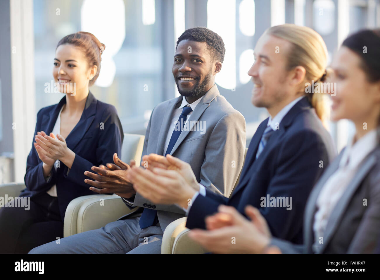 Multi-gruppo etnico di sorridere business persone sedute in fila nella moderna hall di vetro e battimani, focus su allegro imprenditore africana guardando camer Foto Stock