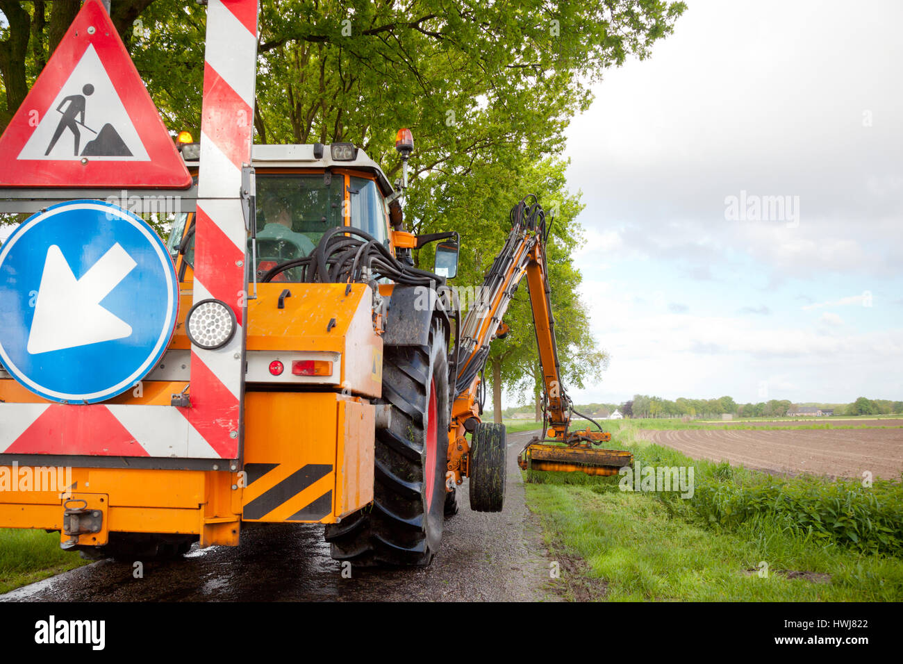 La falciatura di erba spallamento lungo la strada in uno spazio pubblico con grande arancione falciatrice trattore Foto Stock