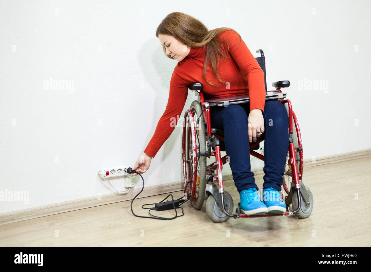 Disabili donna caucasici in carrozzella si allunga per presa di alimentazione a muro per accendere il computer portatile Foto Stock