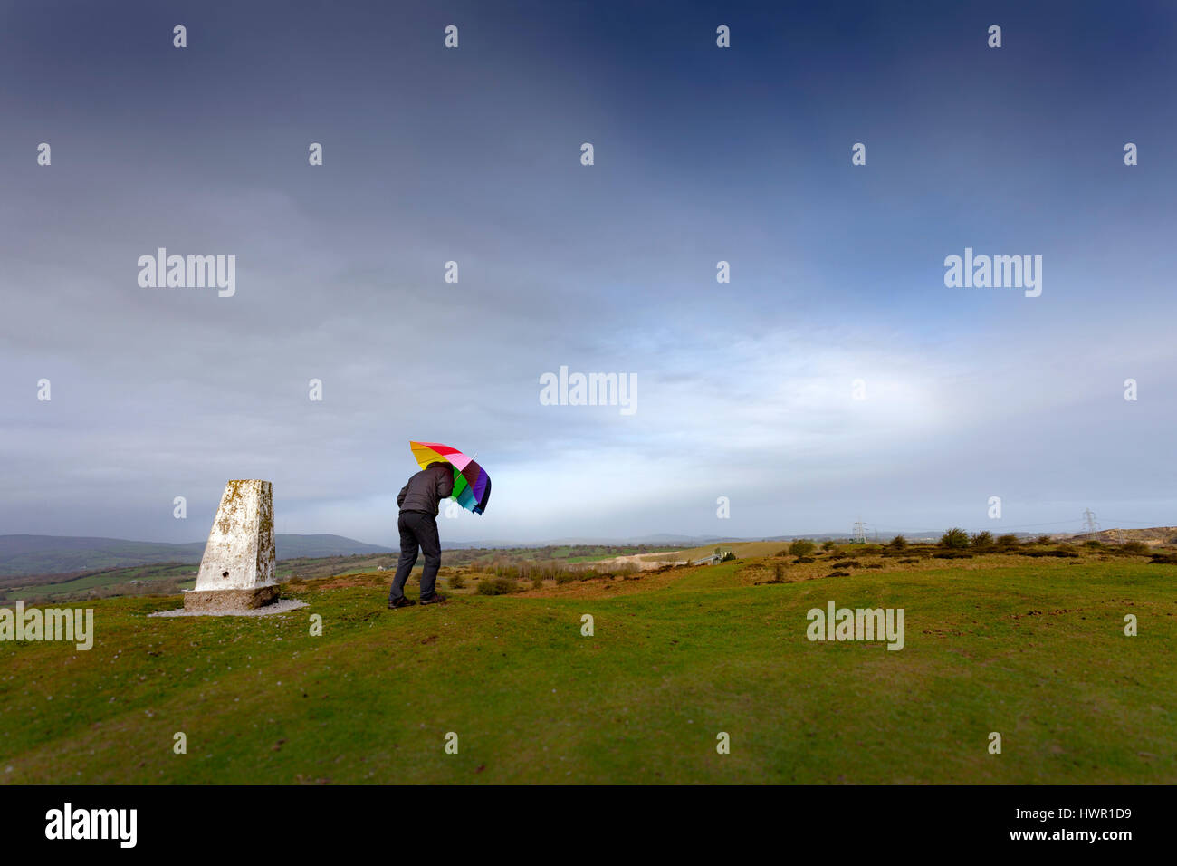 Persona in possesso di ombrello contro il vento accanto ad un pilastro trig sulla Halkyn Montagna in Flintshire, Galles Foto Stock