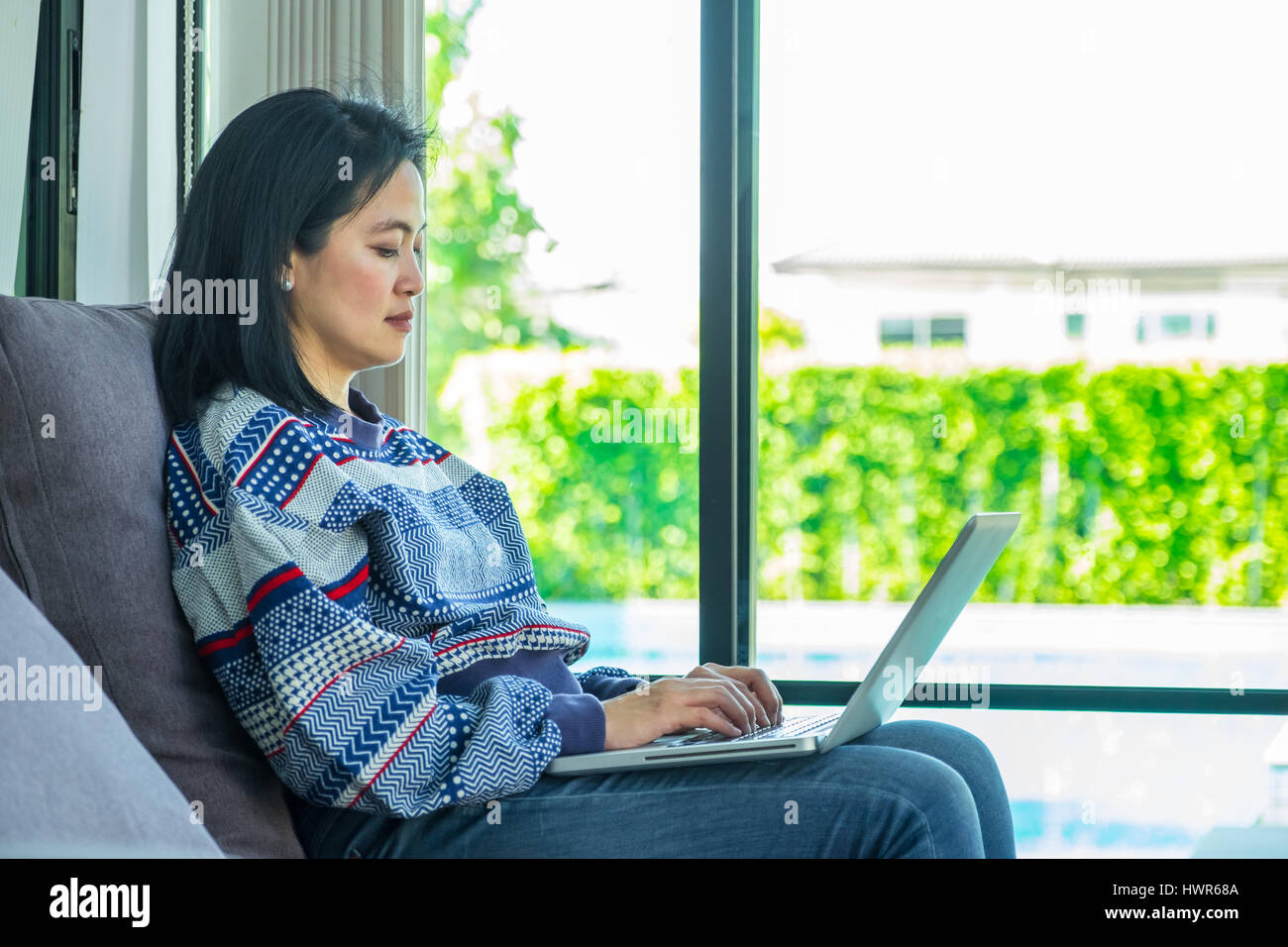 Felice donna seduta su un comodo divano utilizzando laptop finestra vicino a casa,uno stile di vita digitale. Foto Stock