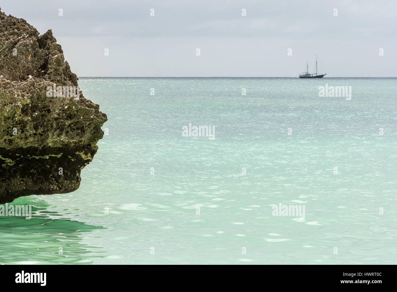 Una linea di riva grande roccia appendere fuori su una luce verde paradiso mare con una grande barca a vela off all'orizzonte. Foto Stock