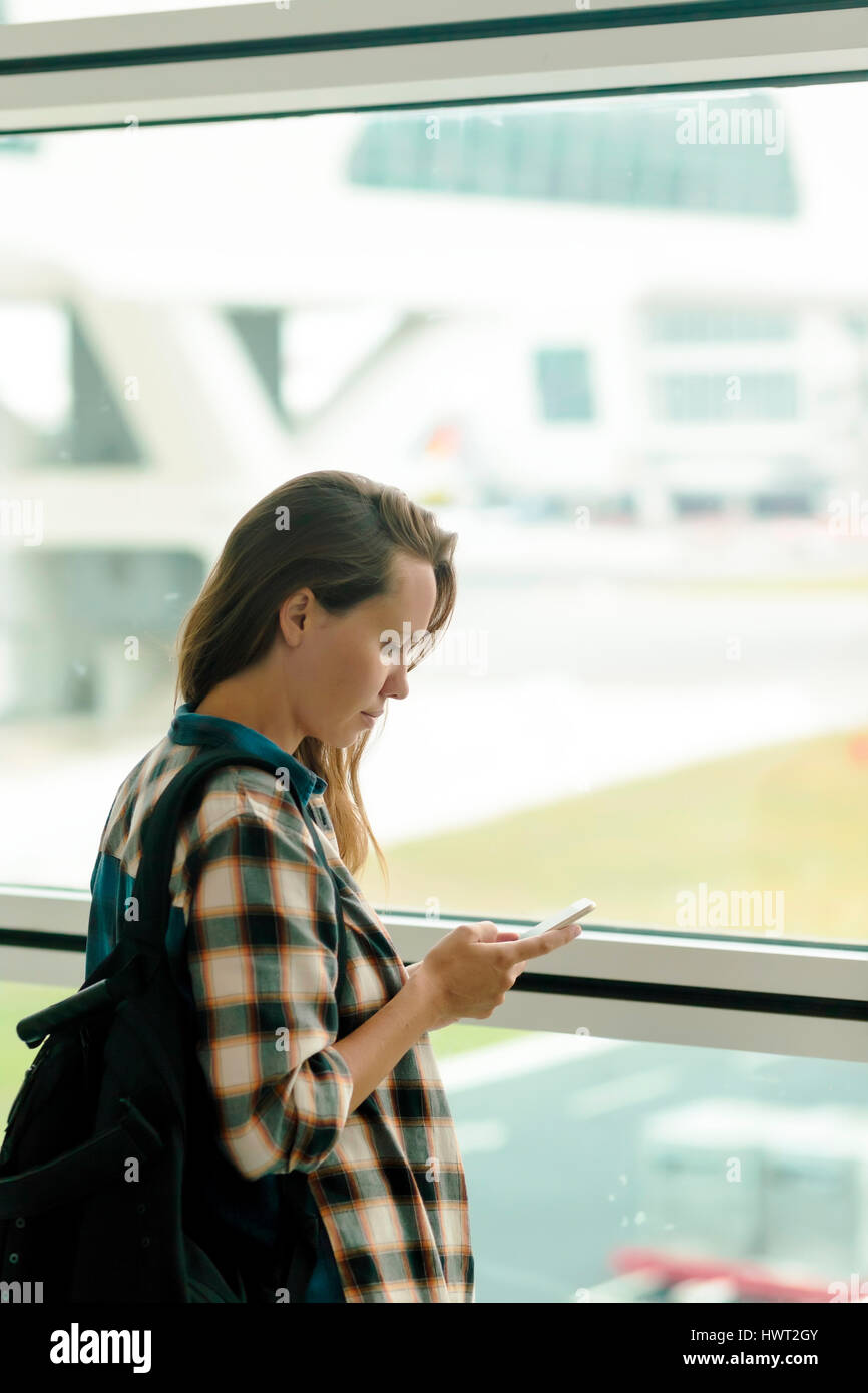 Donna che utilizza il cellulare mentre in piedi da una finestra in aeroporto area di partenza Foto Stock