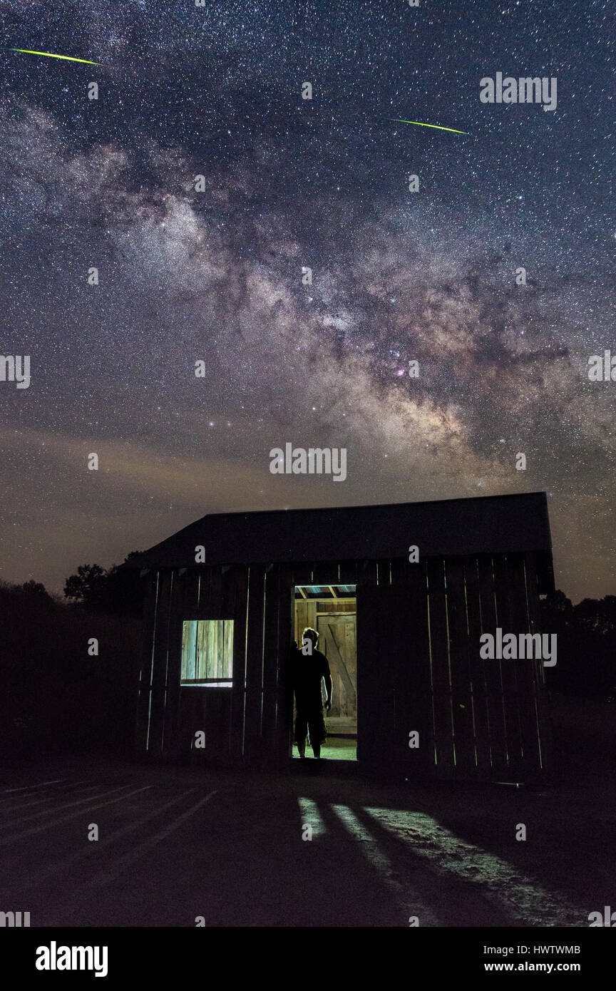 Un uomo si stagliano contro un back lit lonely capannone sotto un drammatico cielo notturno con la via lattea sopra. Foto Stock