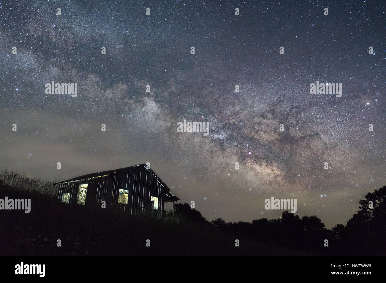 Le stelle e la Via Lattea nucleo galattico, separate da un sottile velo di nebbia, incombono su di un paesaggio scuro e un lone shack illuminato dall'interno. Foto Stock