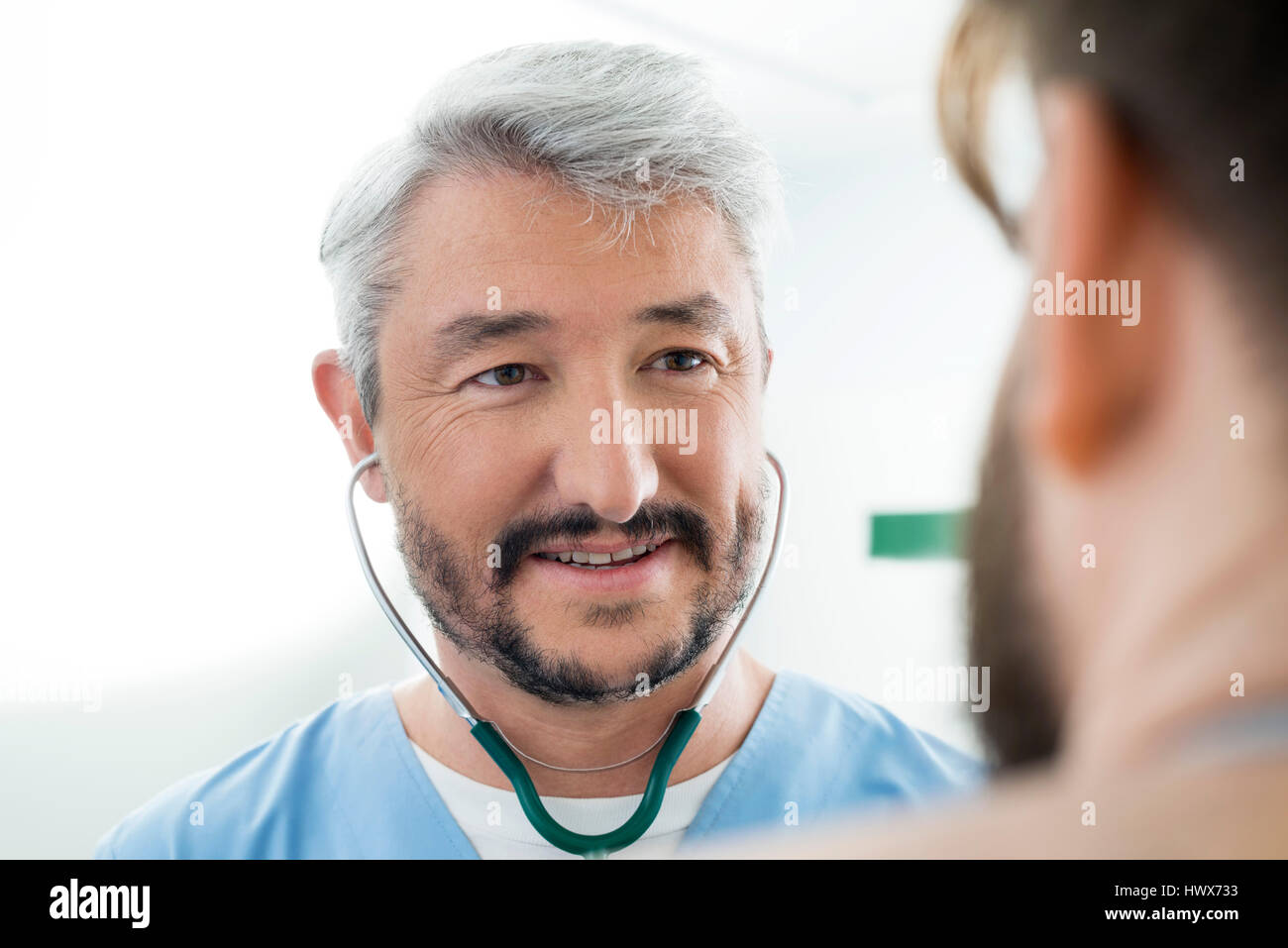 Sorridente medico maschio indossa uno stetoscopio mentre guarda il paziente in clinica Foto Stock