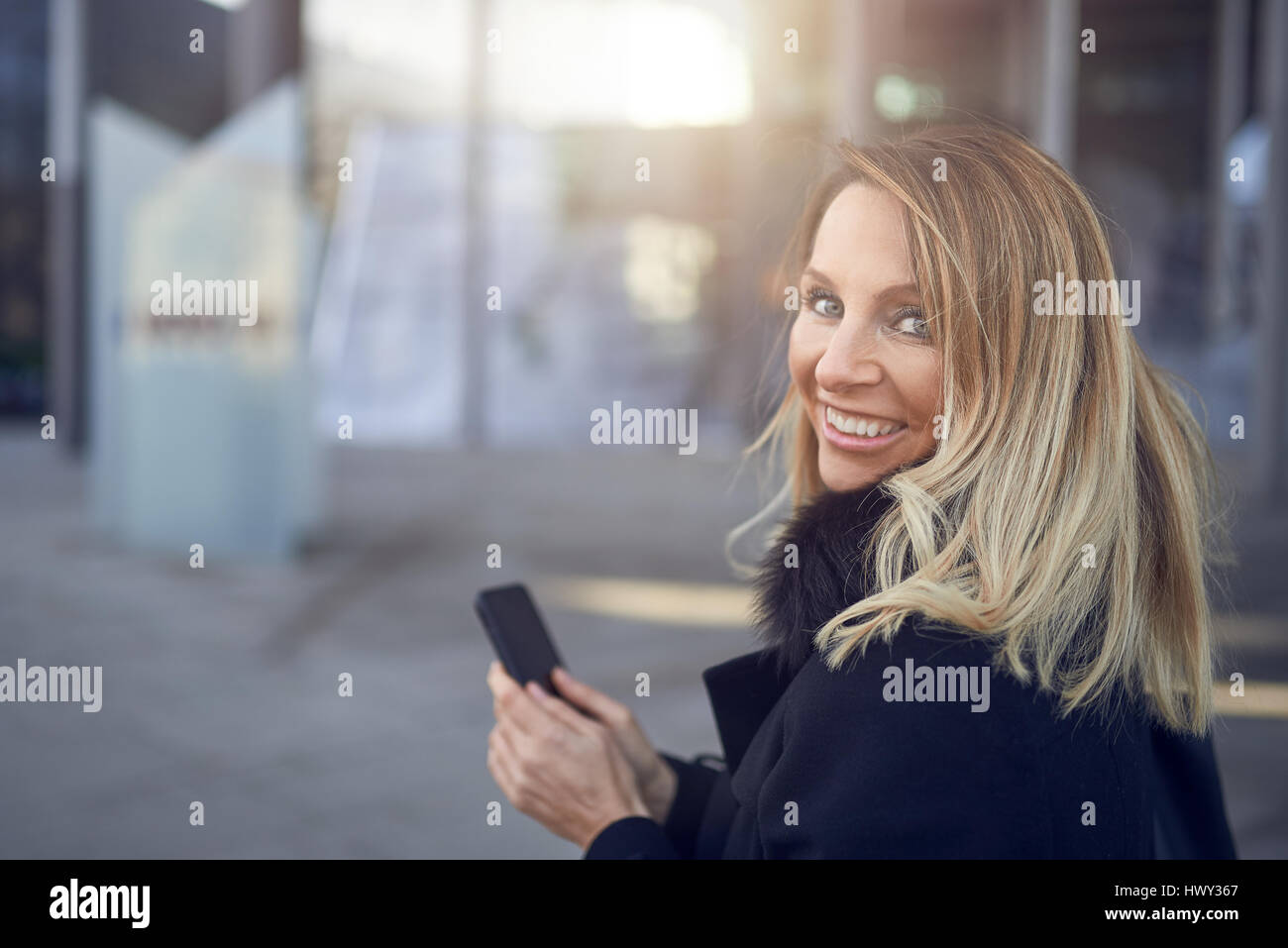 Sorridente donna attraente guardare indietro sopra la sua spalla come essa si erge in una strada della città in possesso di un telefono cellulare con spazio di copia Foto Stock
