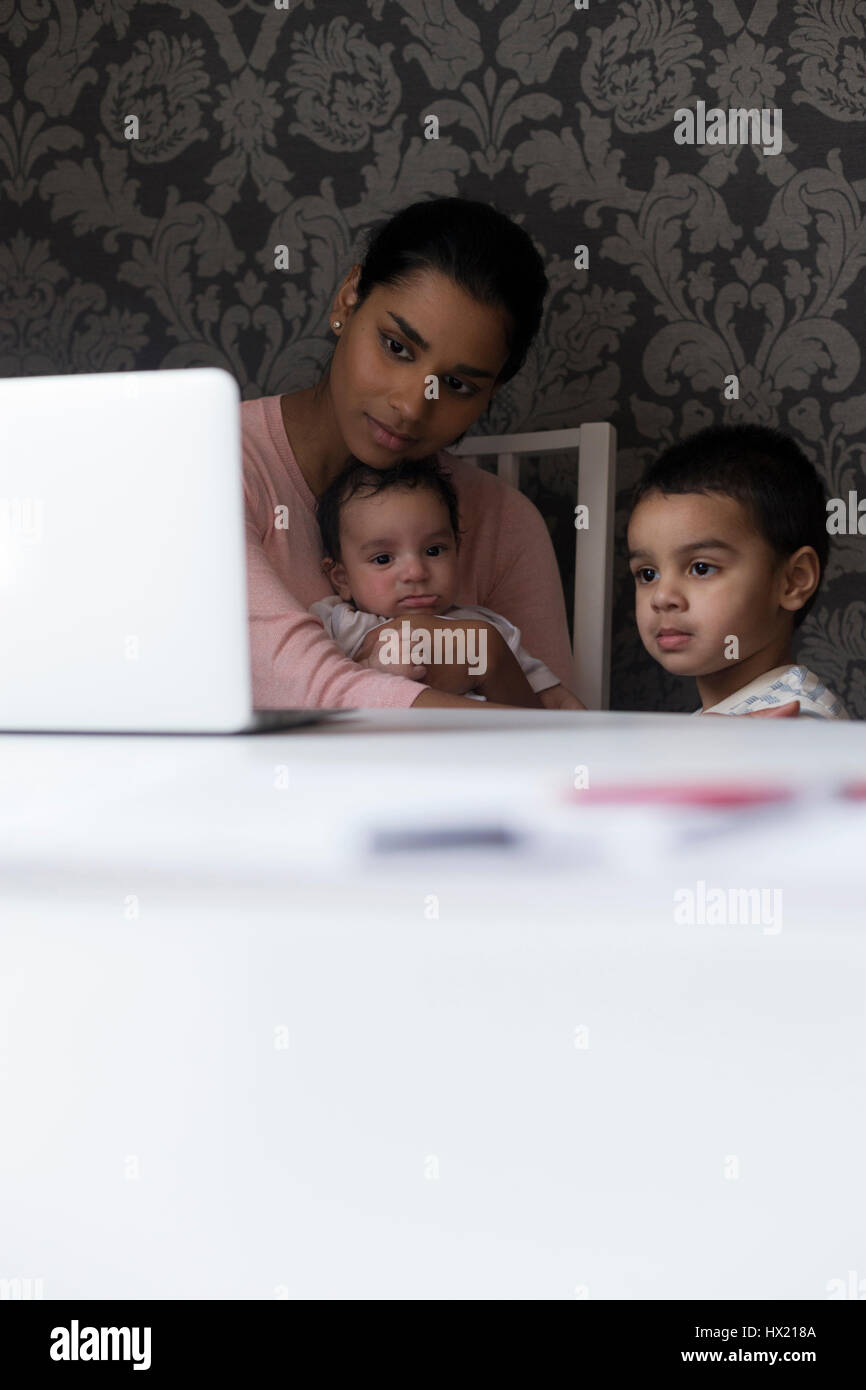 Giovane madre mantenendo i suoi bambini intrattenuti da mostrare loro qualcosa sul suo computer portatile. Lei tiene il suo bambino figlio e il primogenito è in piedi accanto a Foto Stock