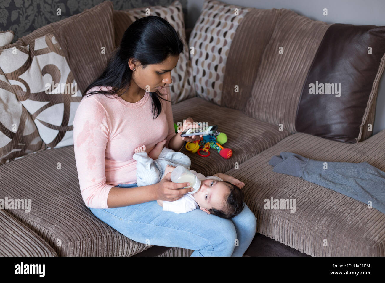 Giovane madre con il suo smartphone mentre alimentando il suo bambino figlio di una bottiglia di latte. Foto Stock