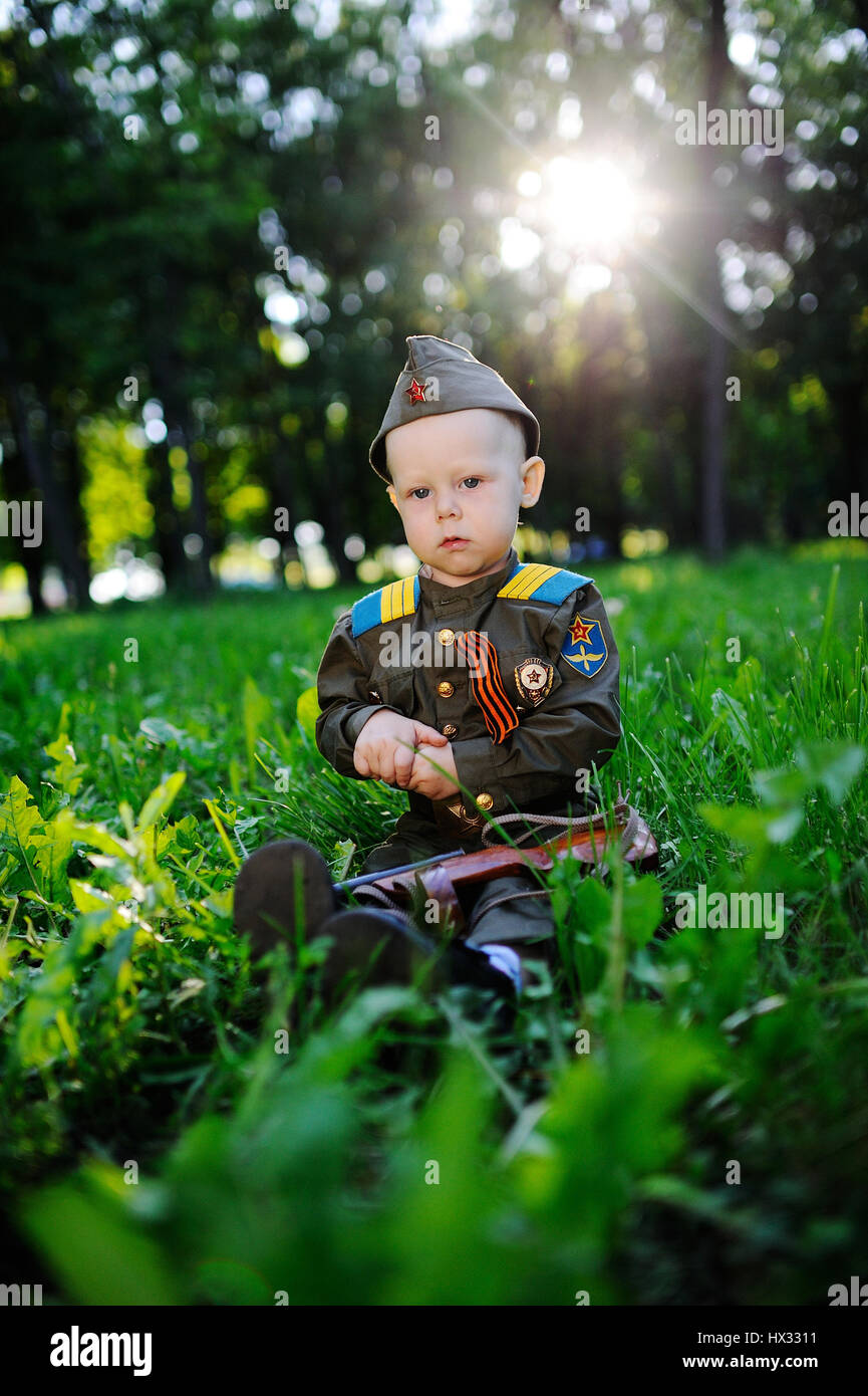 Il bambino in uniforme militare con un nastro di assedio si siede in erba contro il tramonto. Il 9 maggio la Giornata della Vittoria, 23 febbraio Foto Stock