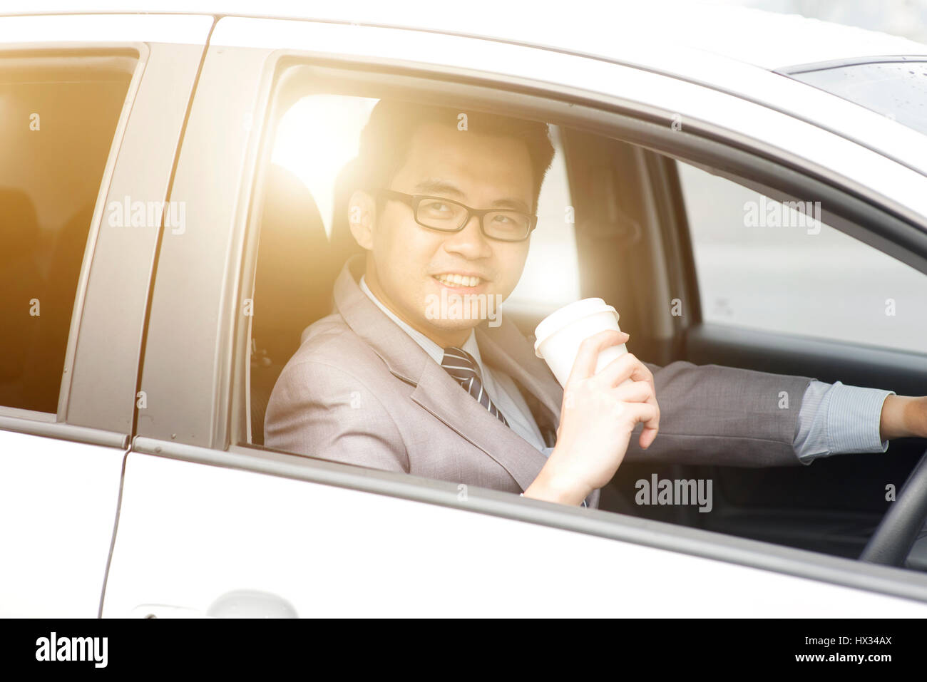 Asian business uomo alla guida di una vettura e tenendo la carta tazza di caffè al mattino con la splendida luce del sole. Foto Stock