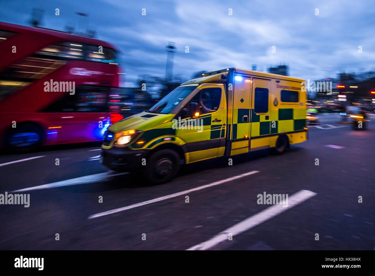 Un'ambulanza che viaggia ad alta velocità al crepuscolo con luci blu che lampeggiano a Londra e che passa davanti a un autobus rosso Foto Stock