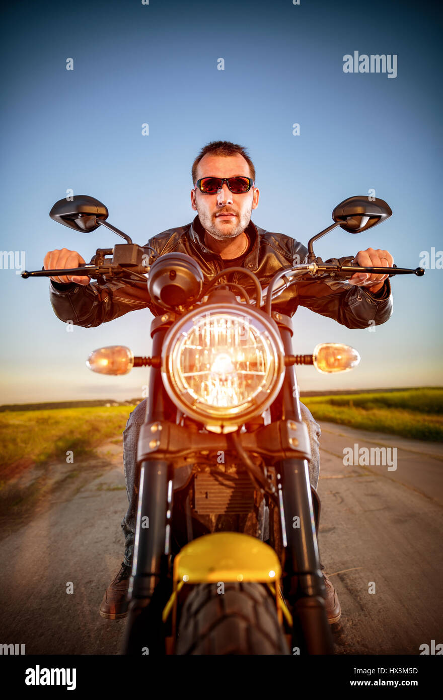 Biker uomo che indossa una giacca di pelle e occhiali da sole seduto sulla sua moto guardando il tramonto. Foto Stock
