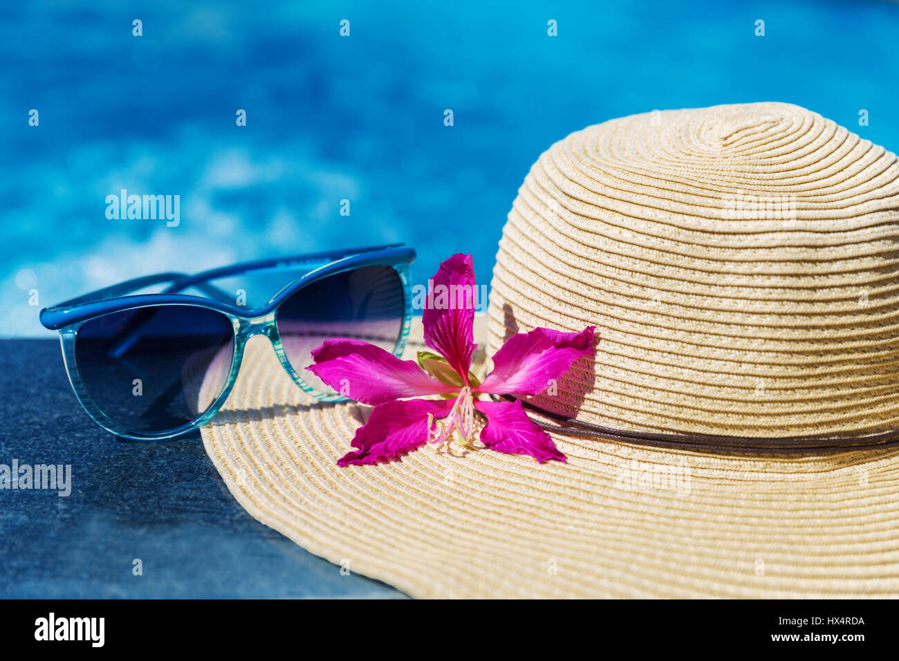 Occhiali da sole con fiore e cappello di paglia sul bordo di una piscina - vacanza concetto tropicale Foto Stock