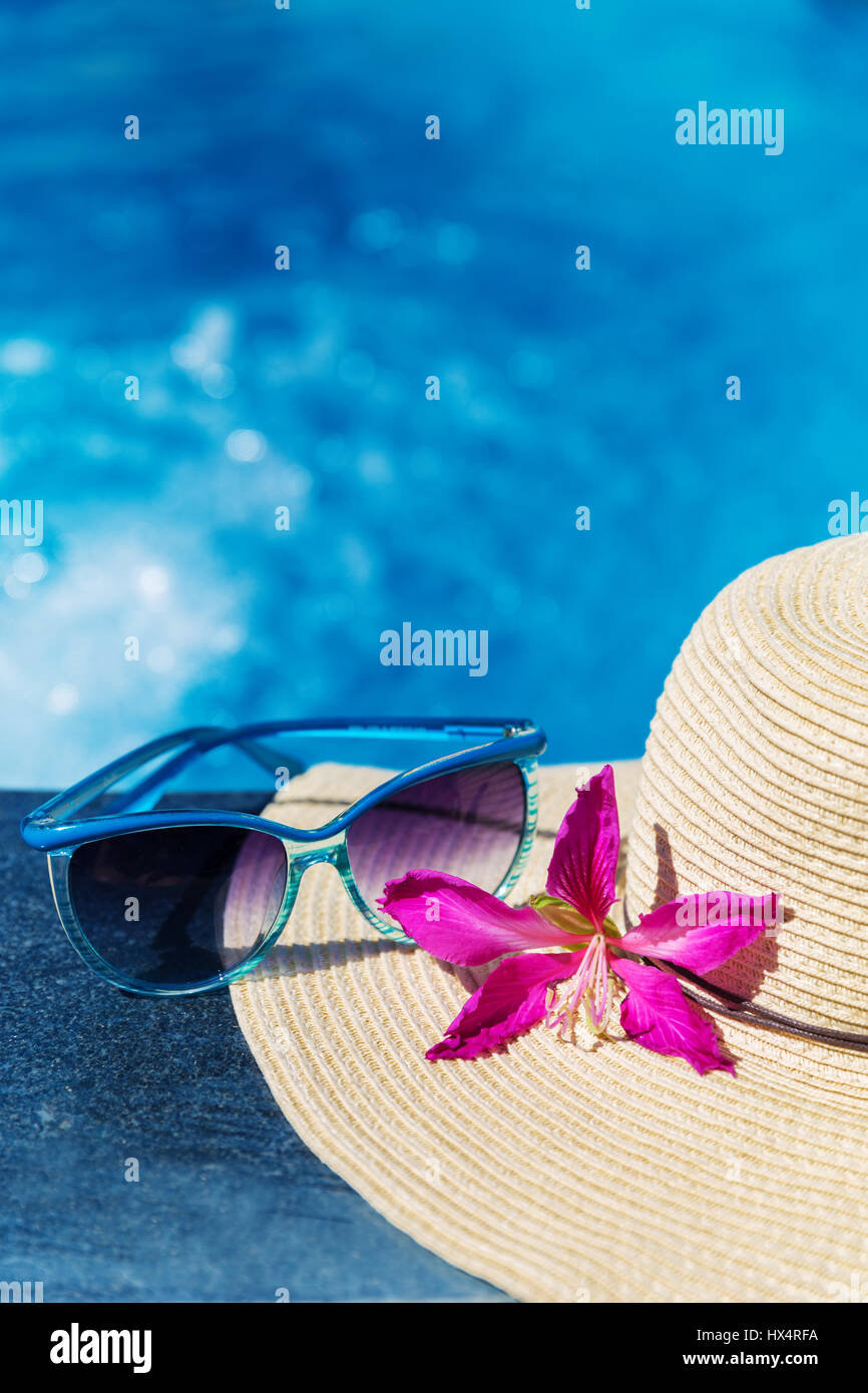 Occhiali da sole con fiore e cappello di paglia sul bordo di una piscina - vacanza concetto tropicale Foto Stock