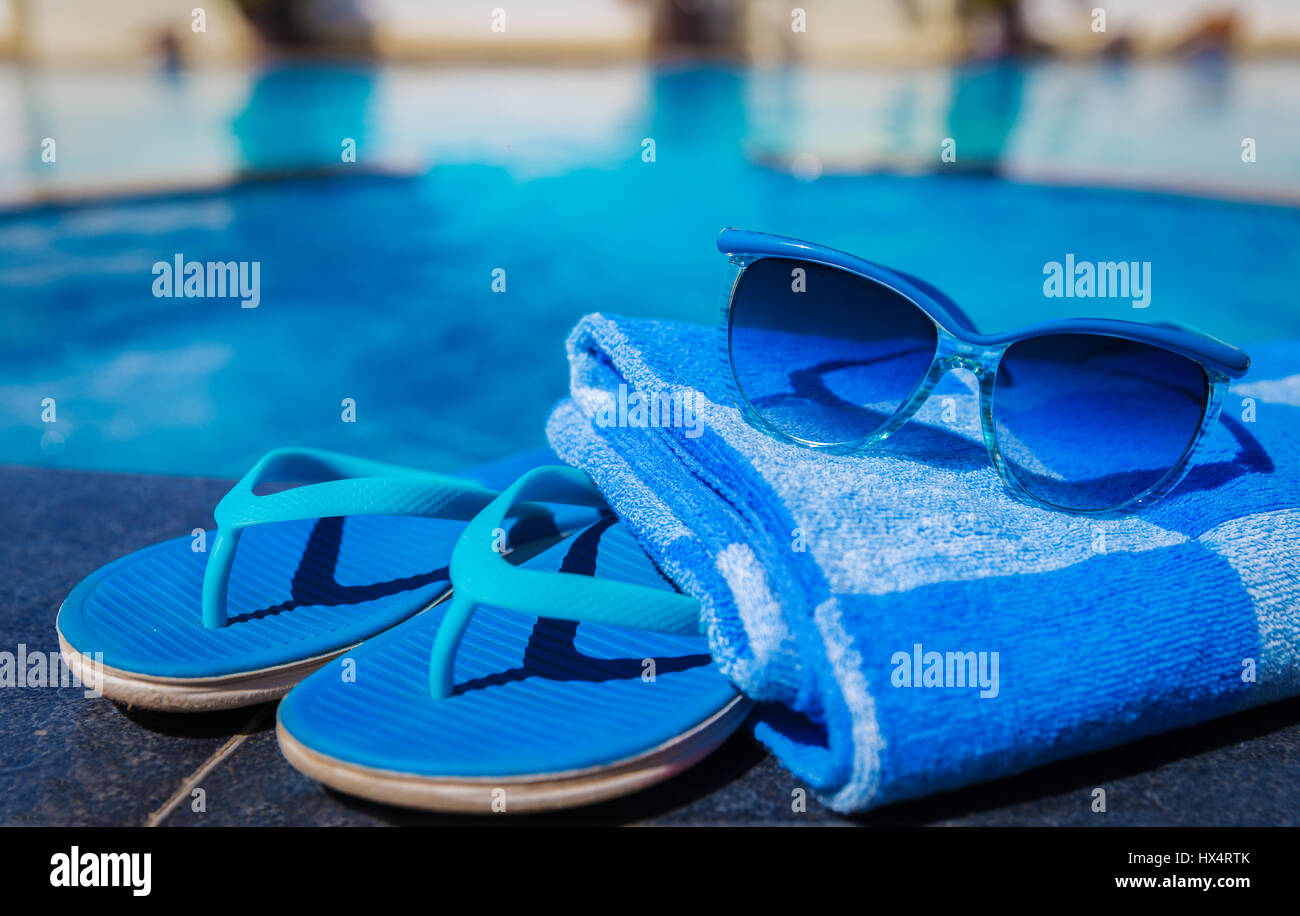 Blue pantofole, occhiali da sole e la salvietta sul bordo di una piscina - vacanza concetto tropicale Foto Stock