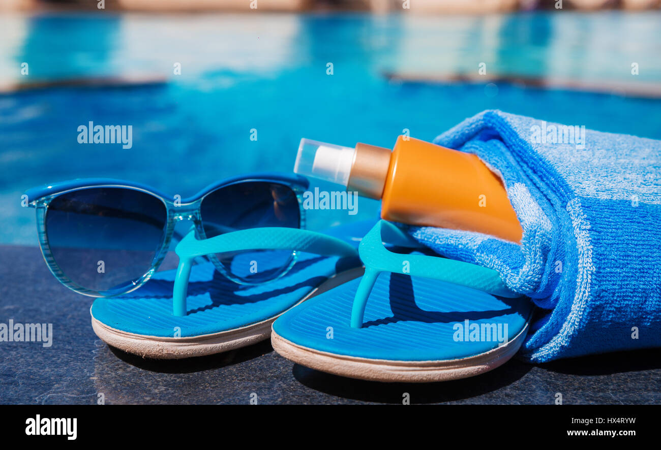 Occhiali da sole con crema di protezione solare, blu pantofole e asciugamano sul bordo di una piscina - vacanza concetto tropicale Foto Stock
