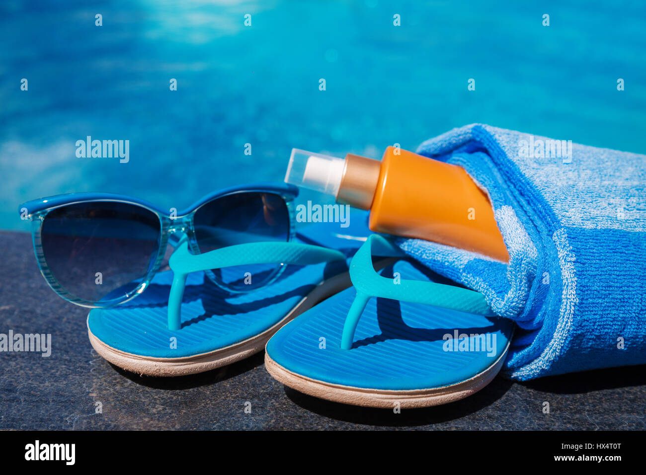 Occhiali da sole con crema di protezione solare, blu pantofole e asciugamano sul bordo di una piscina - vacanza concetto tropicale Foto Stock